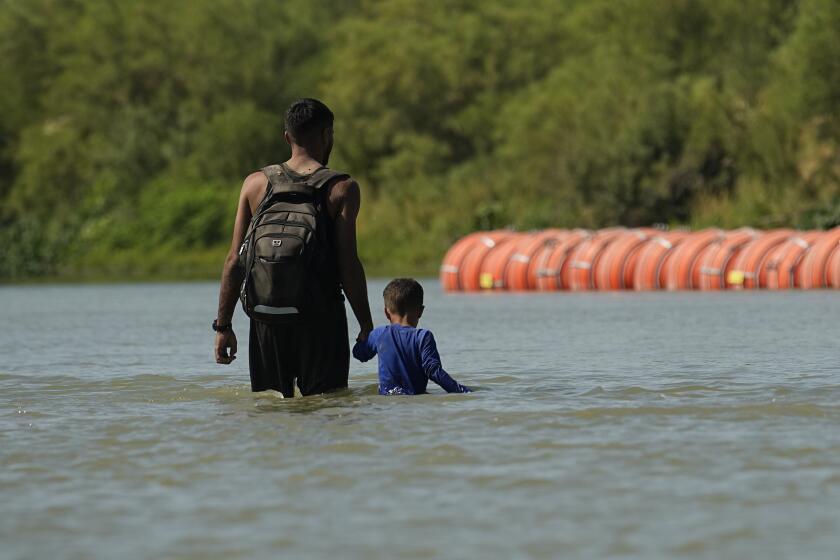 ARCHIVO - Dos migrantes caminan cerca de unas grandes boyas usadas como barrera fronteriza flotante en el Río Grande, o Bravo, el 1 de agosto de 2023, en Eagle Pass, Texas.. (AP Foto/Eric Gay, Archivo)