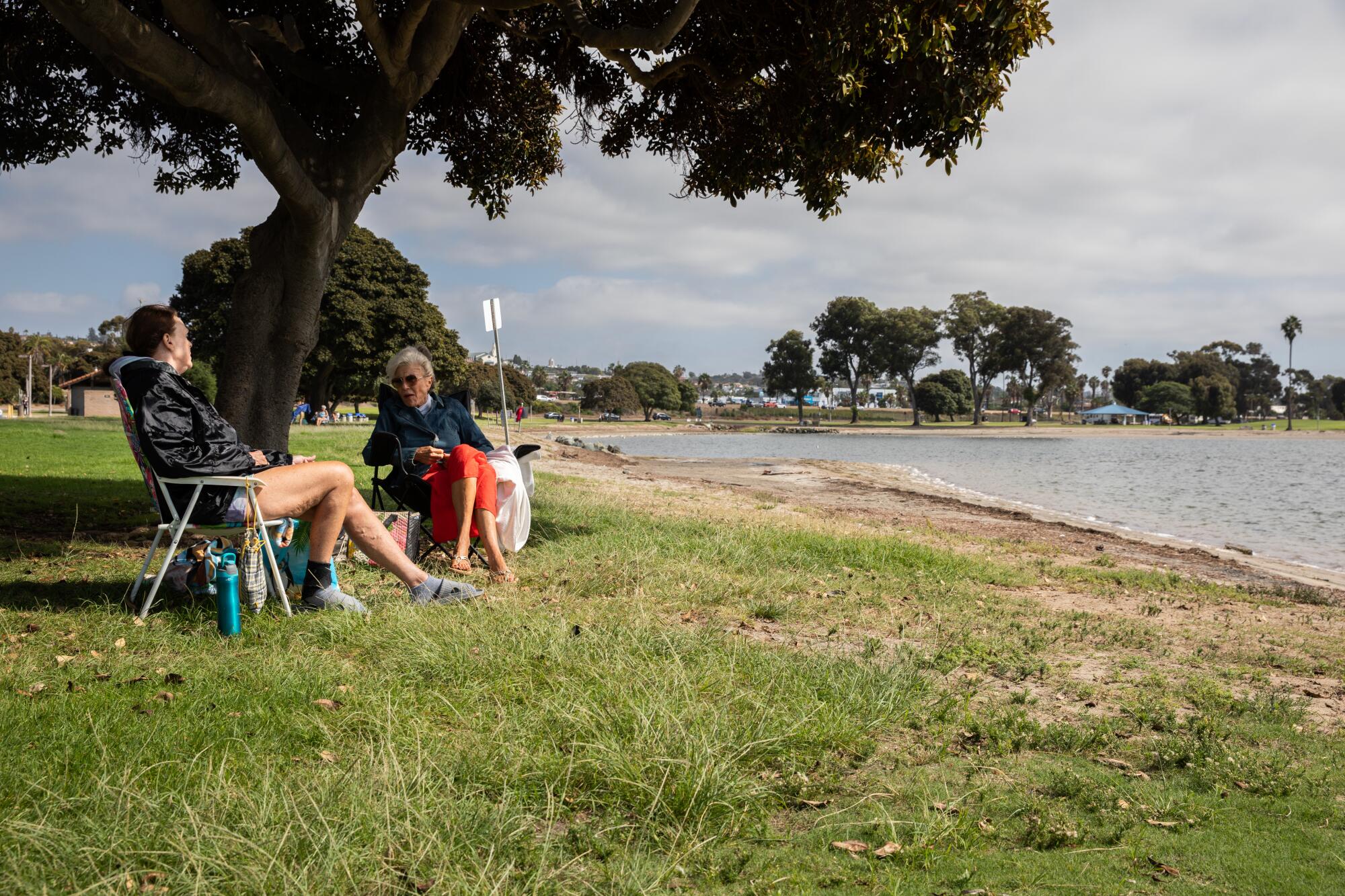 Two women sit on folding chairs on the grass beneath the shade of a tree next to a bay.