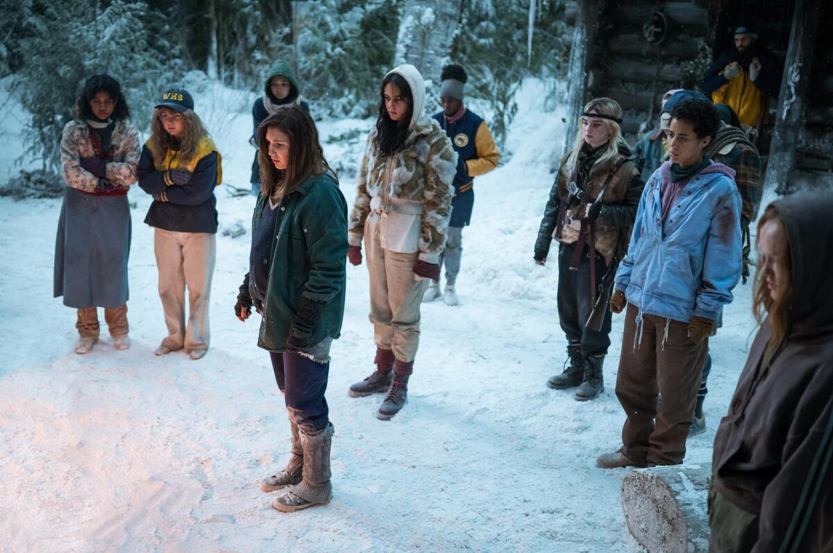 A group of teenage girls stands in the snow, most looking down at the ground in front of them.