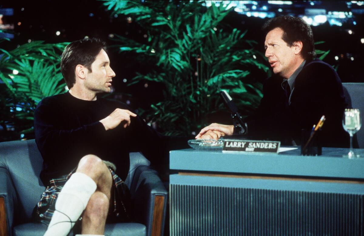 A man wearing a kilt talks to another man sitting behind a desk.