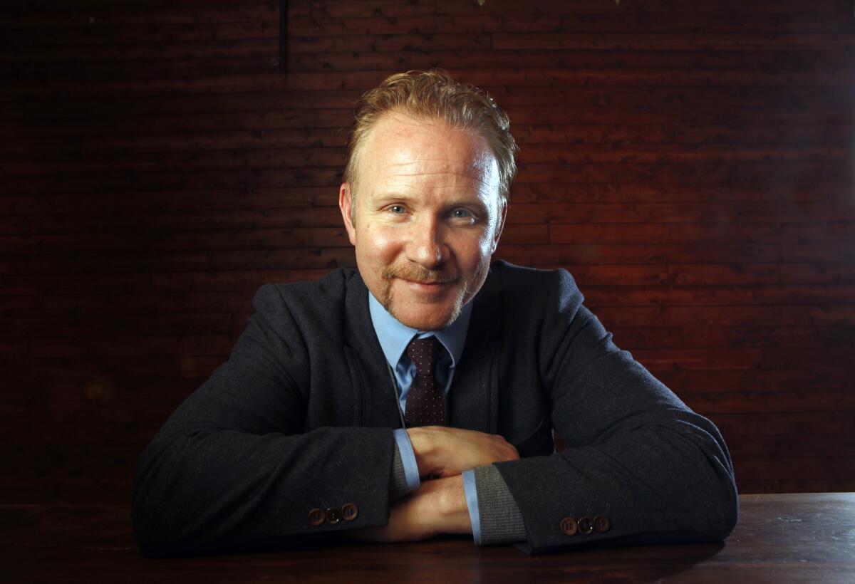 Morgan Spurlock in a dark suit and tie looking straight ahead and smiling slightly with arms crossed on a table