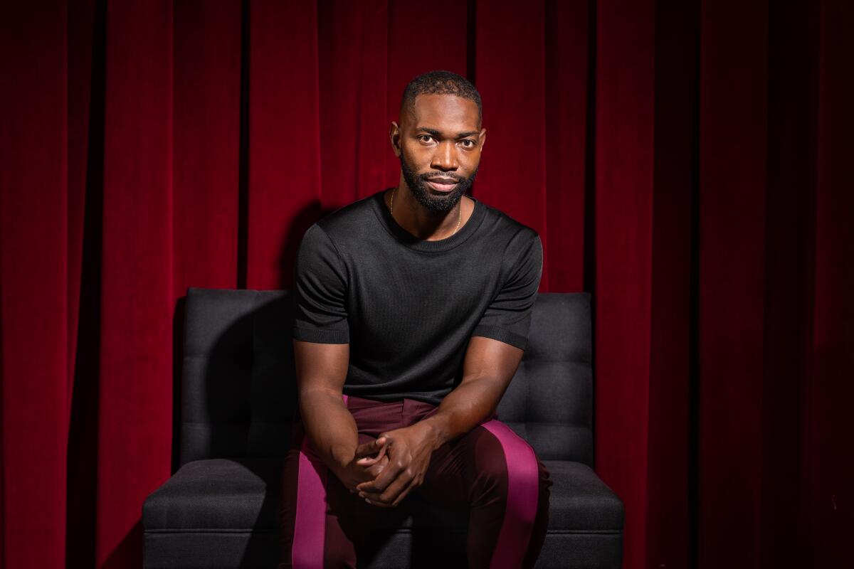 A Black man dressed in black sits in front of red drapes.