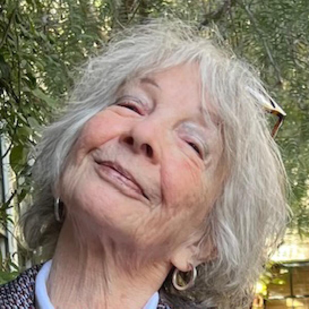 An older woman with short gray hair smiles slightly in a selfie in front of a tree