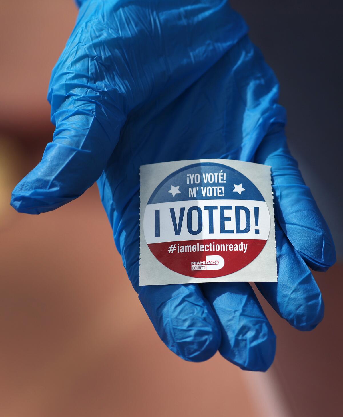 A voter wearing a glove holds an "I voted" sticker. 