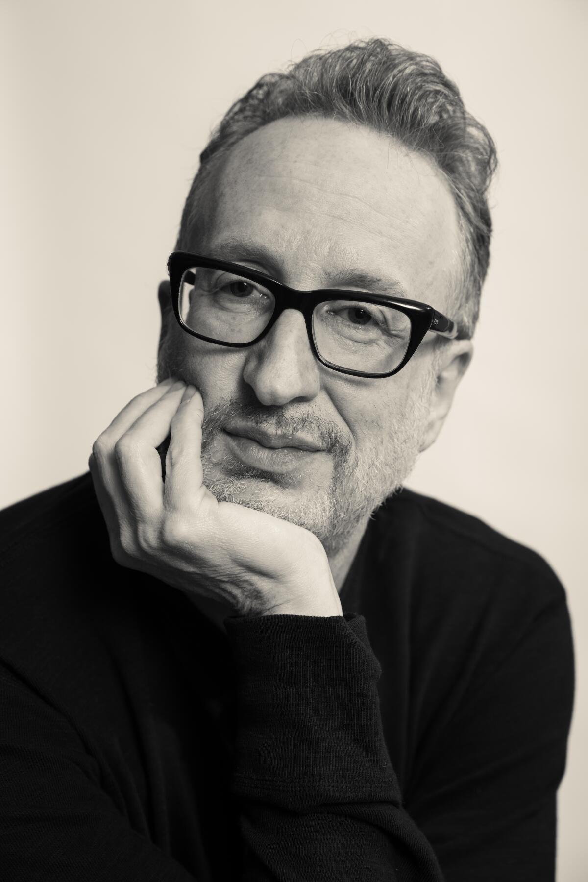 A man in a dark shirt and dark-framed glasses rests his chin on his hand.