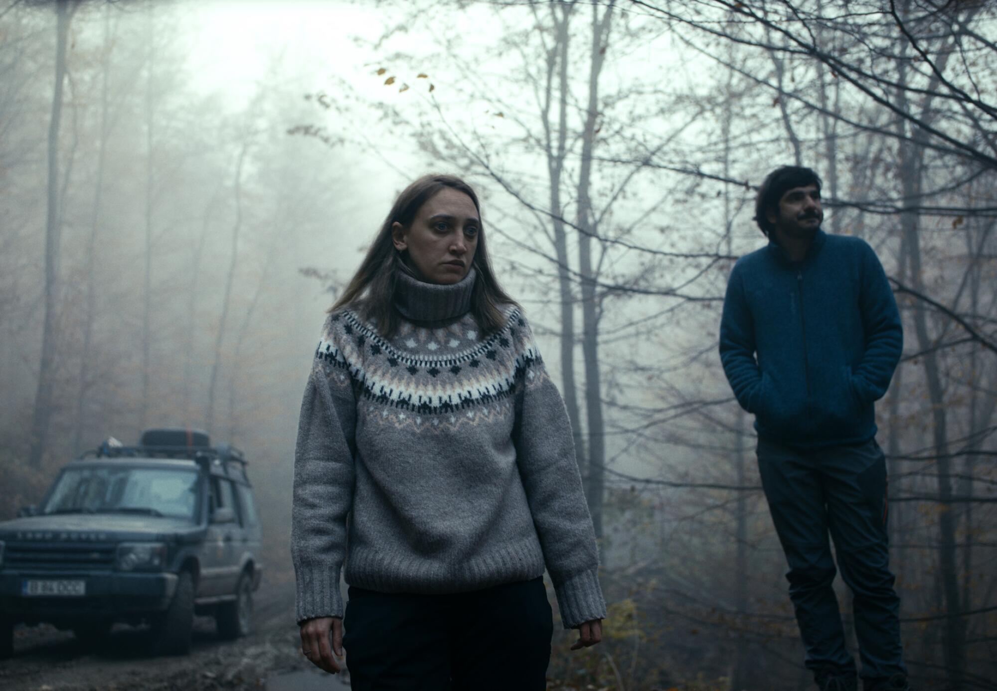 A woman in a chunky sweater and man stand in a foggy forest with a car at their right