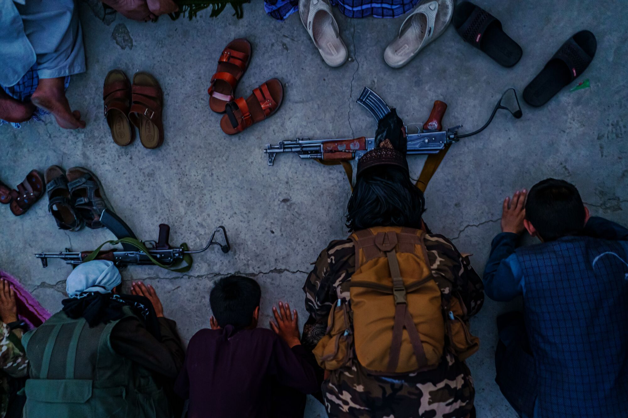 Seen from above, men kneel on the ground, shoes and weapons in front of them.