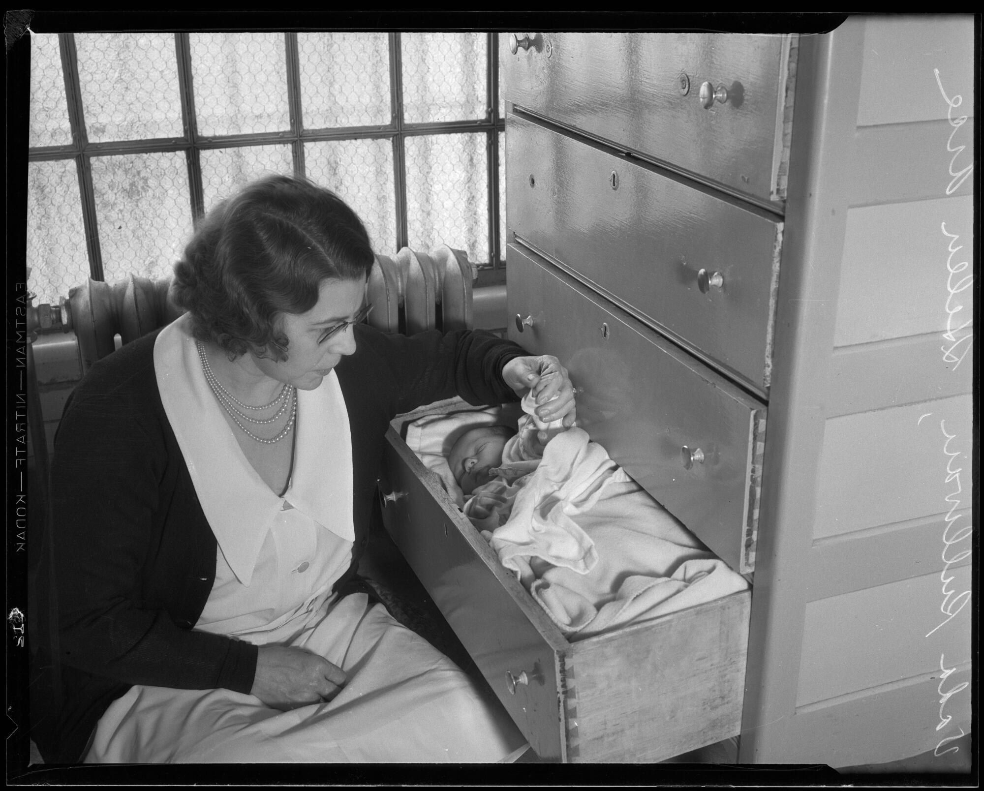 A woman sits next to a dresser with an open bottom drawer serving as a crib for a sleeping baby in 1933.