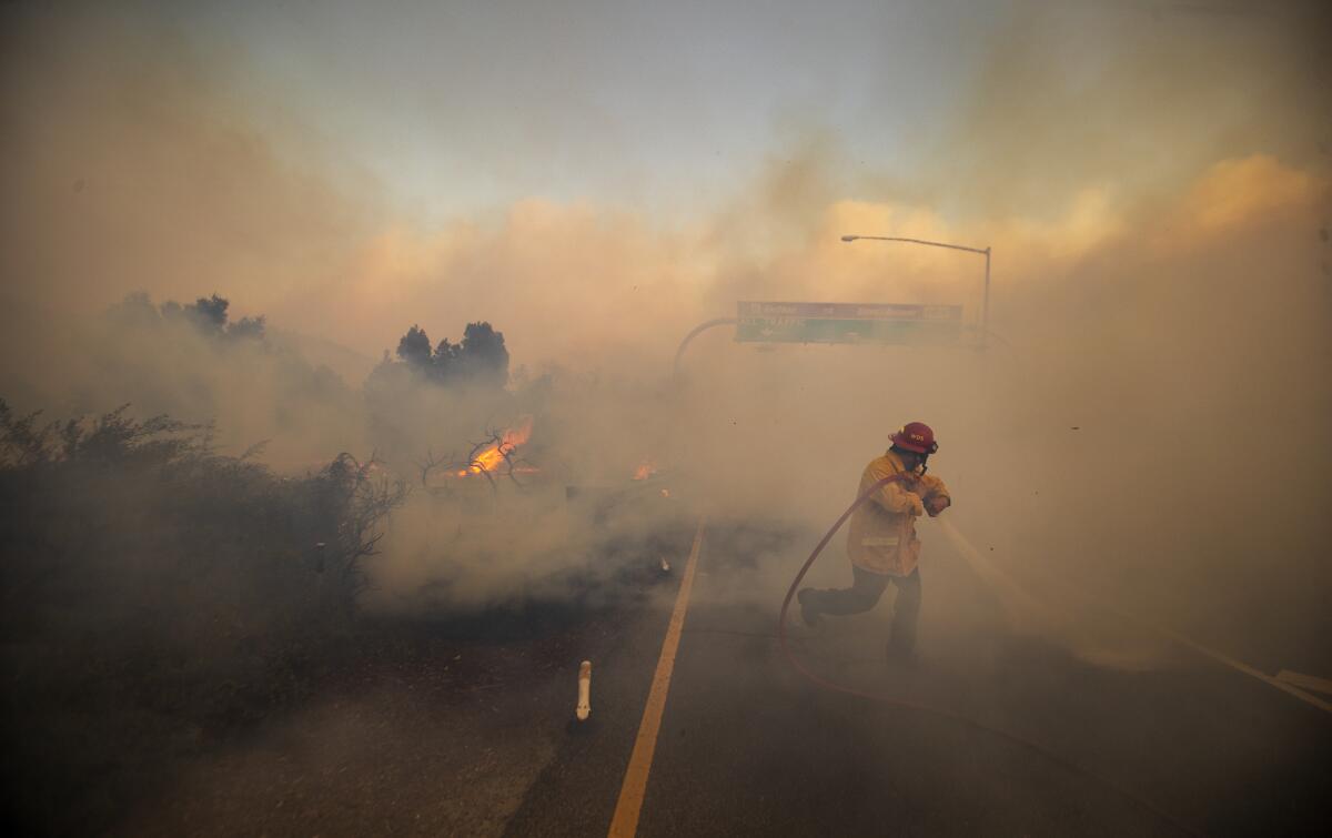 Firefighter Vince Valdivia, standing on a roadway as he sprays a hose, is surrounded by heavy smoke.