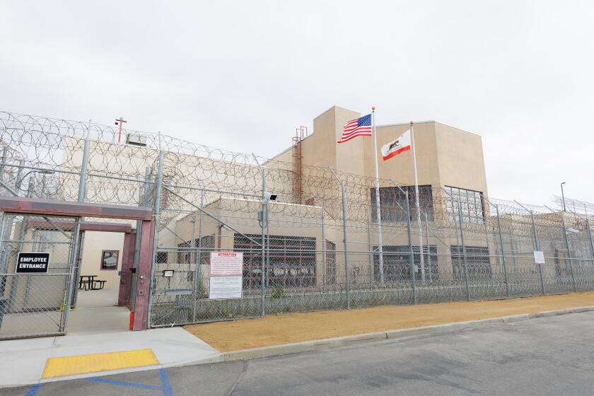 The entrance to a jail is seen behind high barbed wire-topped fencing, with American and California flags flying just inside.