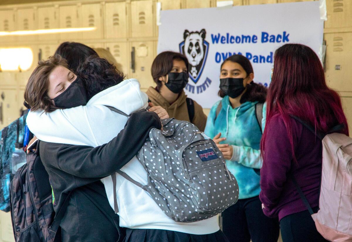 Two girls wearing backpacks hug each other in front of school lockers.