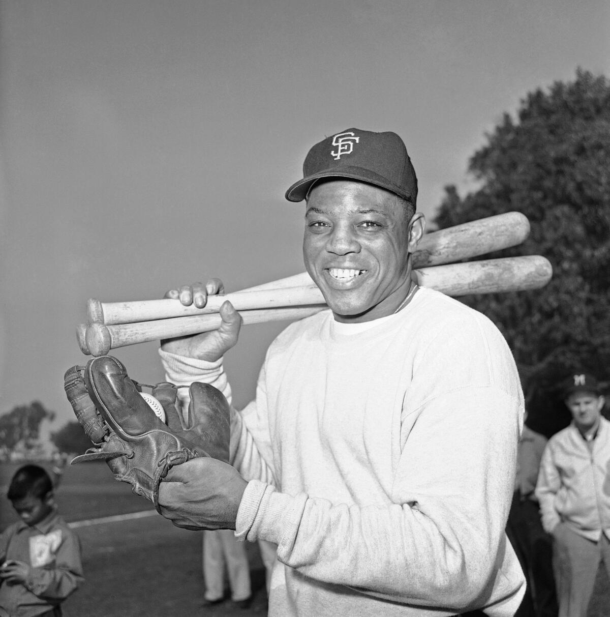 Willie Mays has a baseball glove on one hand and holds several bats in the other while smiling.