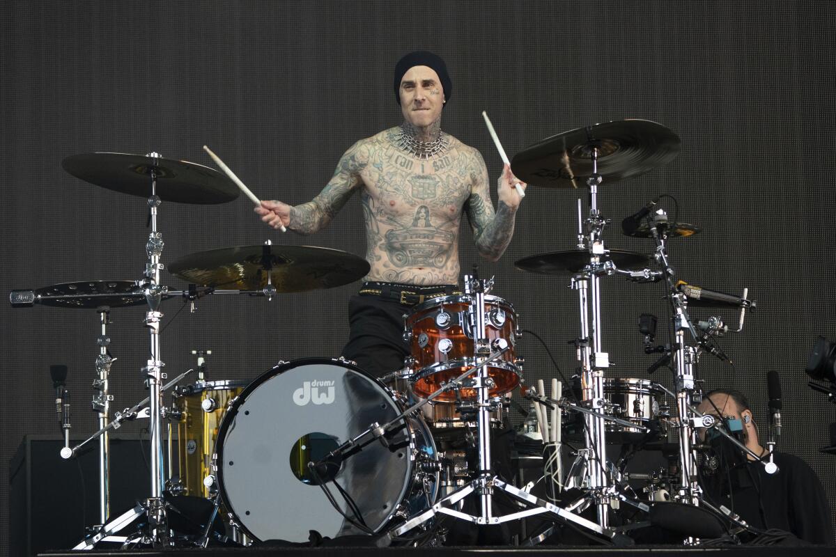 A shirtless, tattooed man in black pants plays a drum set standing up.