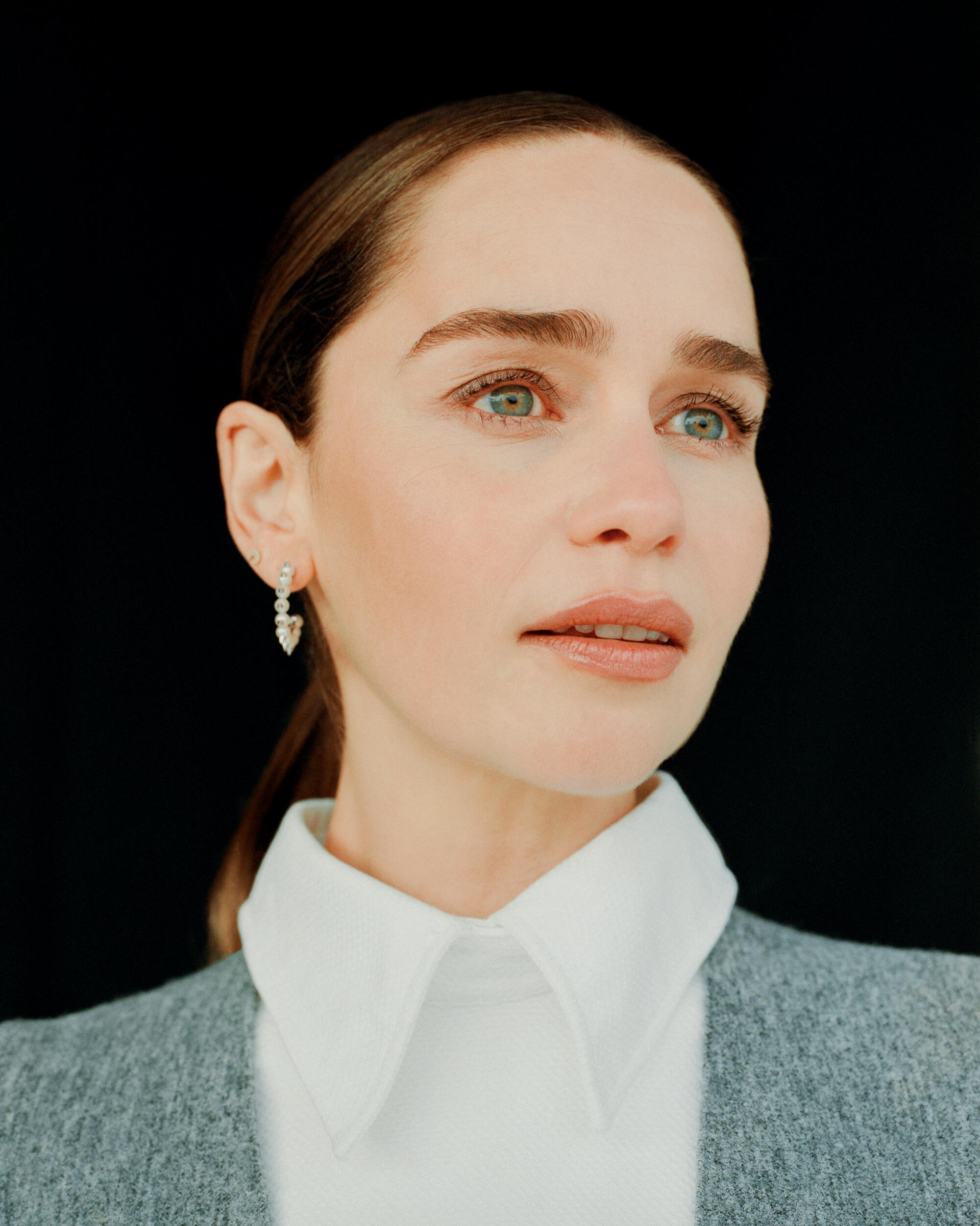 Emilia Clarke, in a collared white shirt and gray jacket, looks to the side.