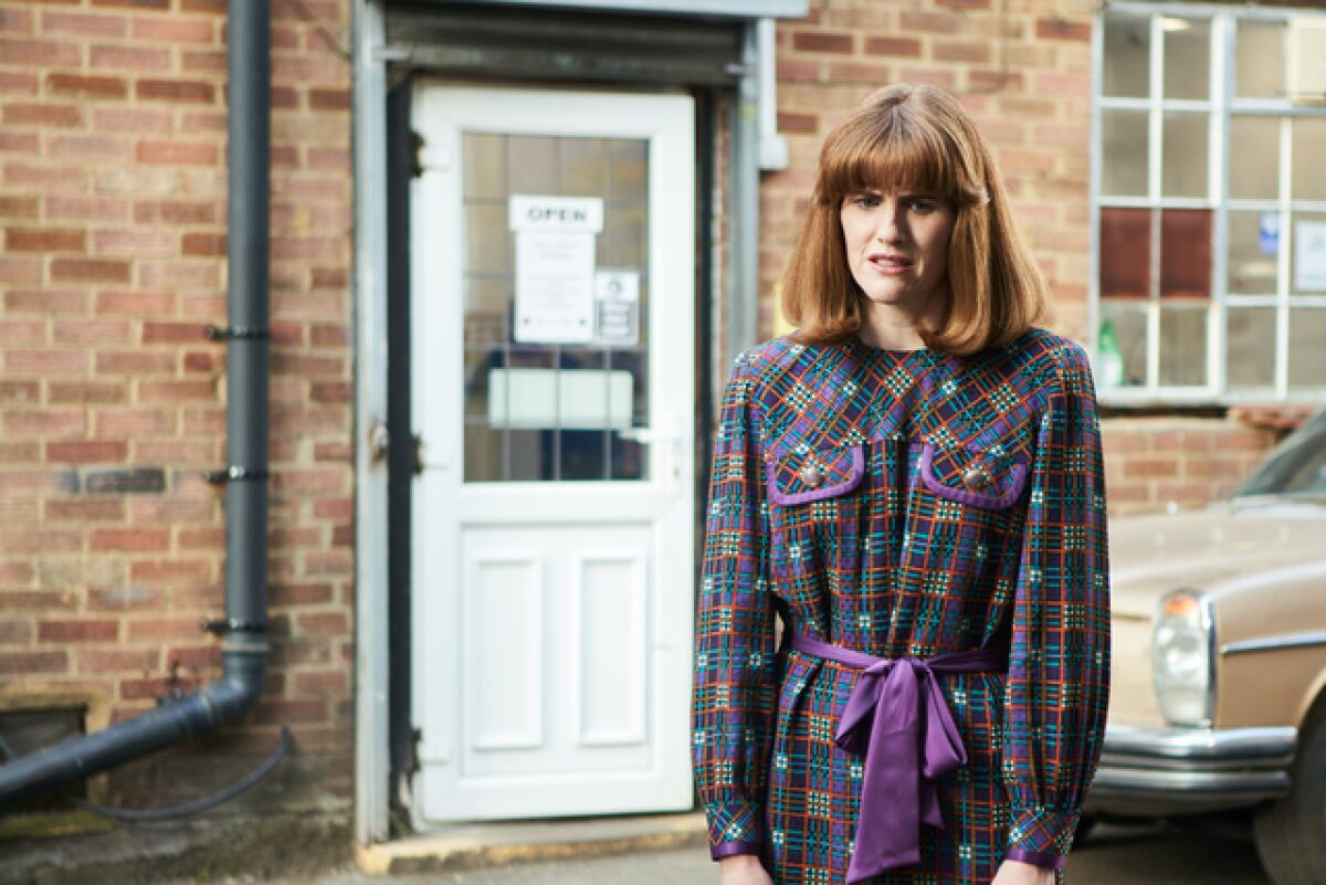 A woman standing outside a brick building in a purple dress looks dismayed.