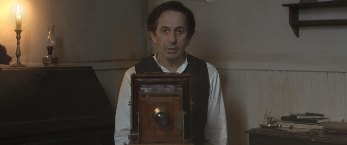 A film still shows an older man standing behind a vintage camera