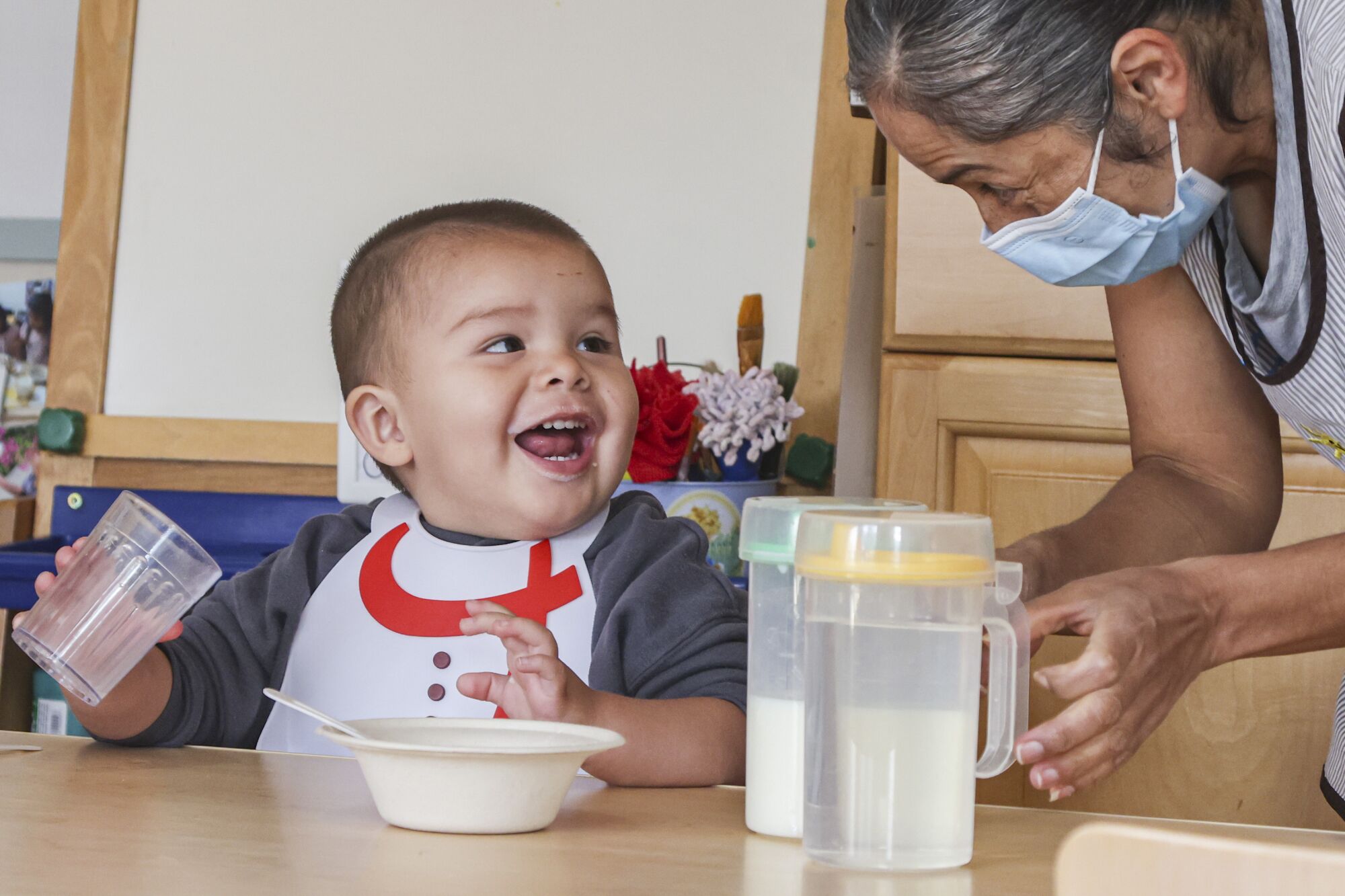 A toddler in a bib holds a cup, laughs and looks up at a woman in a face mask who leans toward him, gesturing playfully.