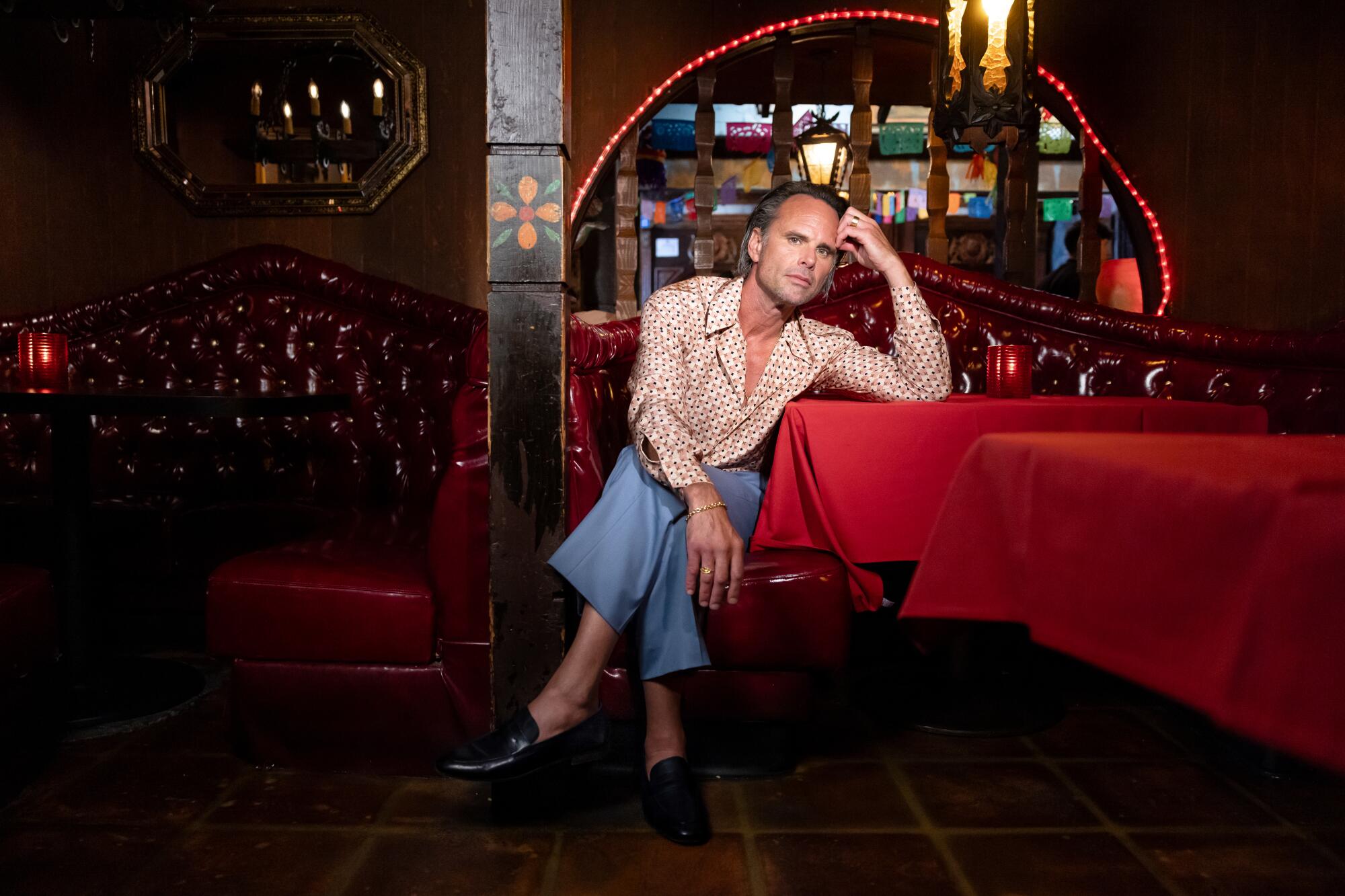 Walton Goggins sits in a red leather booth leaning his arm against a table at a restaurant.