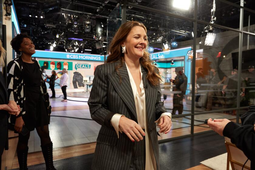 Drew Barrymore is walking off set while wearing a black pinstripe blazer with an off-white shirt and is smiling.