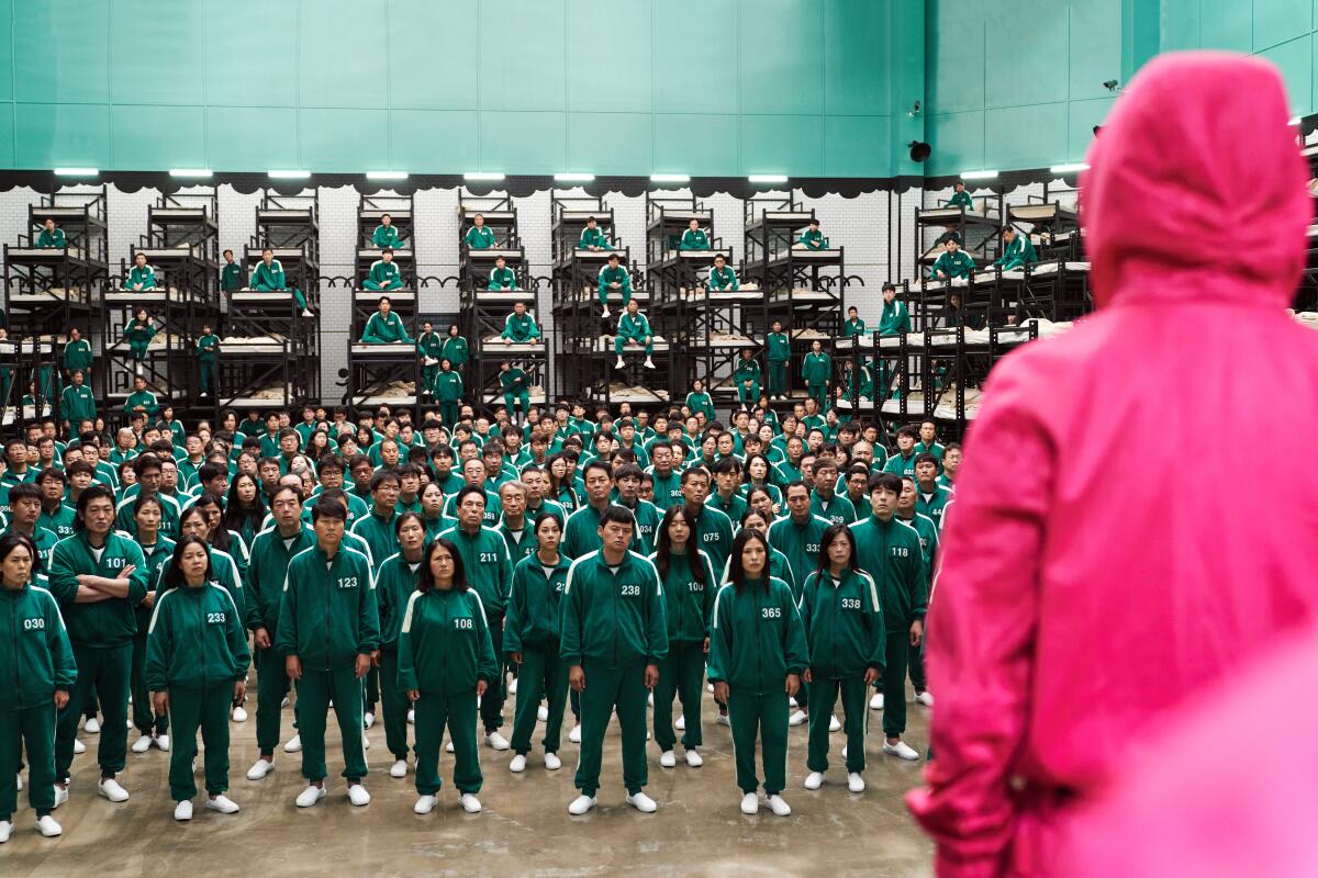 A large group of people in identical green track suits stand in front of a person in a pink hoodie