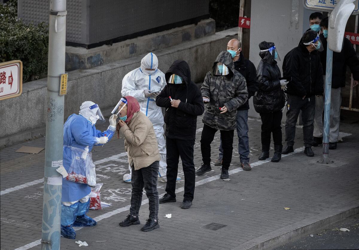 People in masks and face shields line up on asidewalk. A worker in head-to-toe protective gear swabs a person's nose.