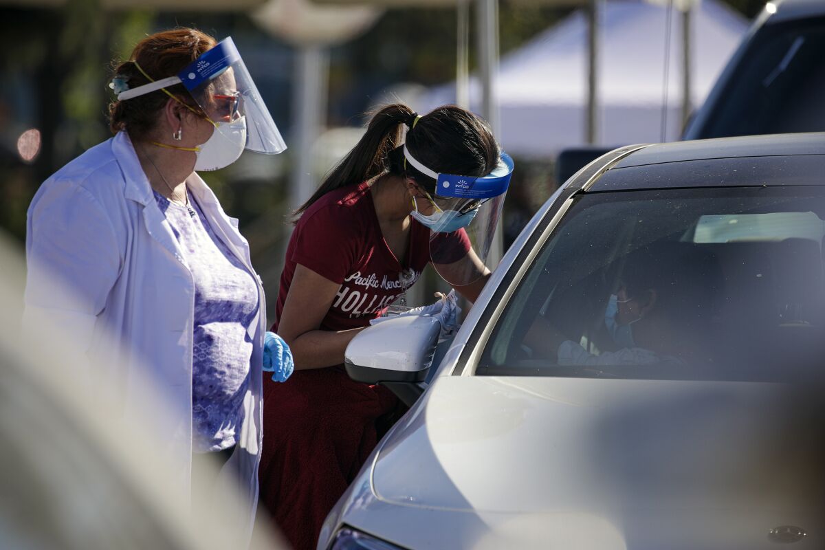 Two health workers with face masks and shields talk to a passenger of a car.