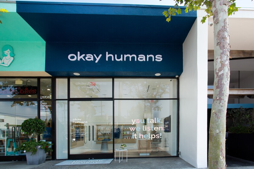 The exterior of a storefront with an "Okay Humans" sign at the top. On the window, it says, "you talk, we listen. it helps!"