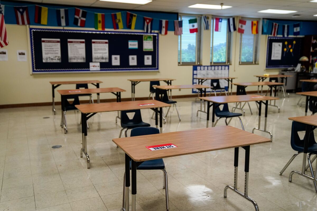 Tables in an empty classroom have "Sit Here" labels to help students with social distancing.