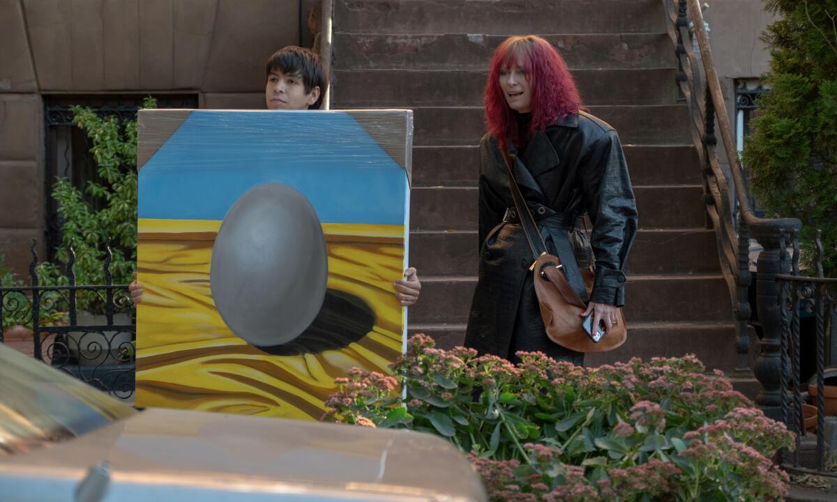 A "Problemista" film still shows Julio Torres holding a painting of an egg next to Tilda Swinton, who wears a red wig
