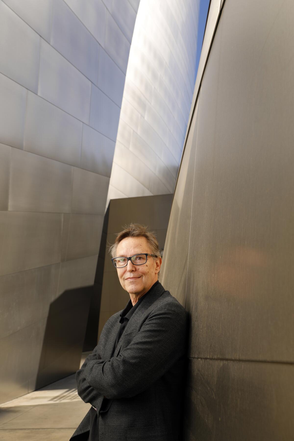 A man in glasses poses against a wall.