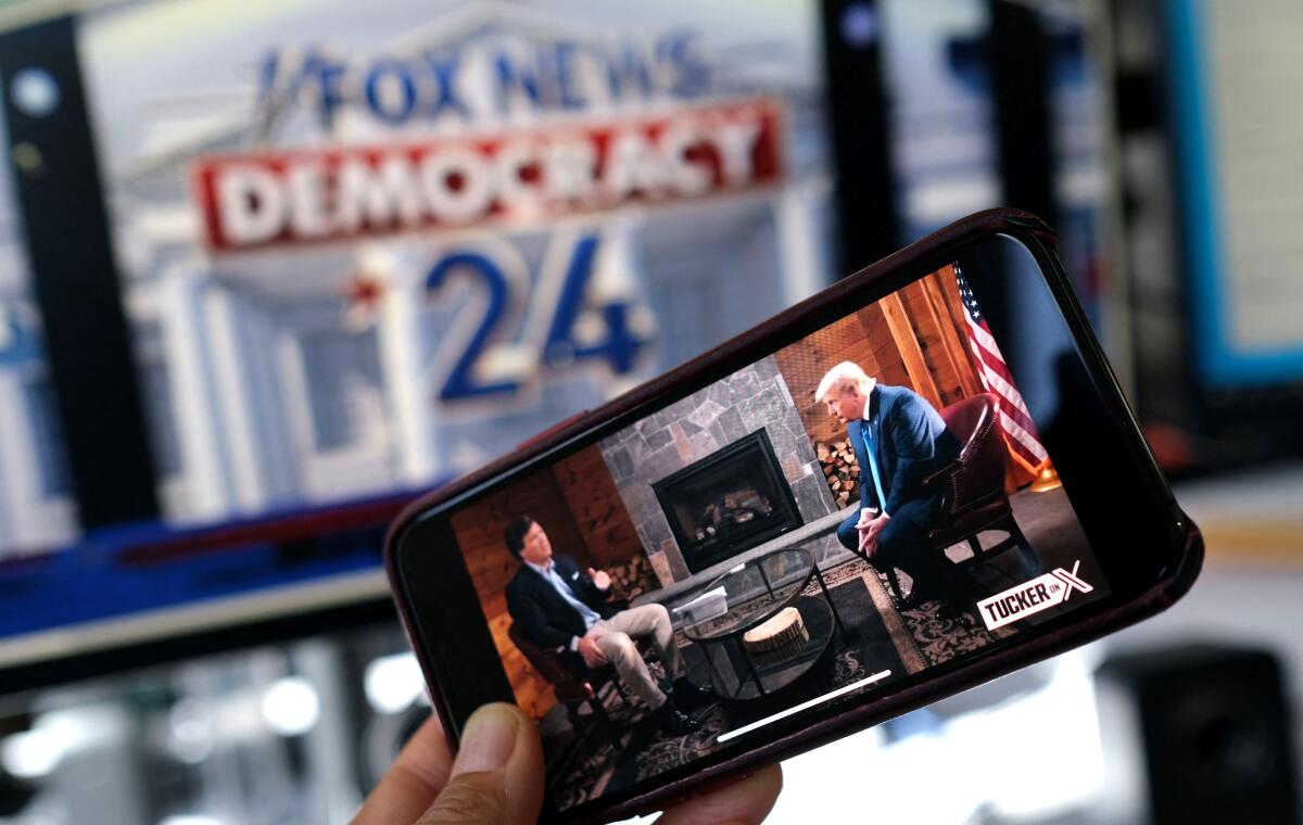 A cellphone with an image of Tucker Carlson and former President Trump is held in front of a screen with Fox News on it.