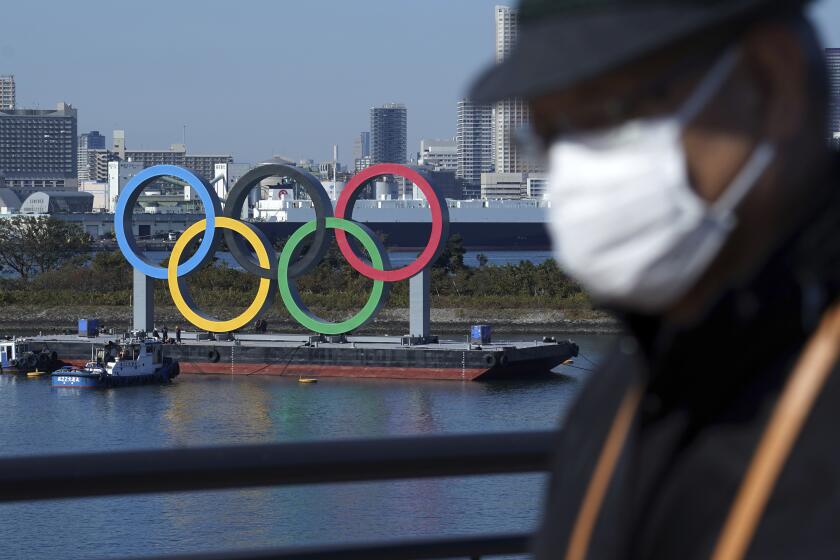 A man wearing a protective face mask to help curb the spread of the coronavirus walks with the Olympic rings in the background in the Odaiba section Tuesday, Dec. 1, 2020, in Tokyo. The rings were removed for maintenance four months ago shortly after the Tokyo Olympics were postponed until next year because of the COVID-19 pandemic. (AP Photo/Eugene Hoshiko)