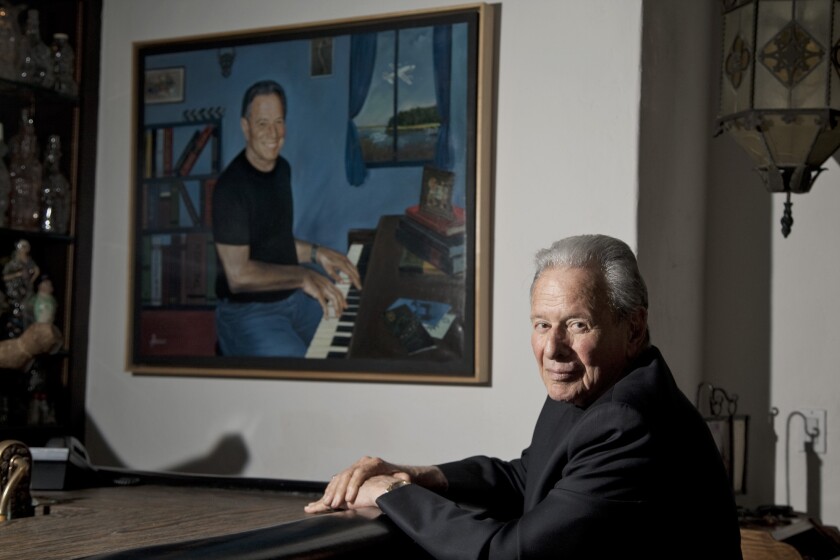 A gray-haired man seated near a painting of a man playing piano.