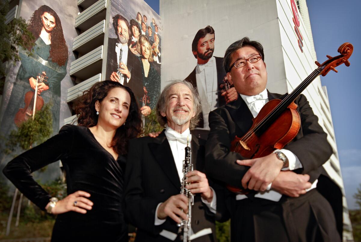 Three musicians, some bearing their instruments, stand before a building-sized L.A. mural bearing their image