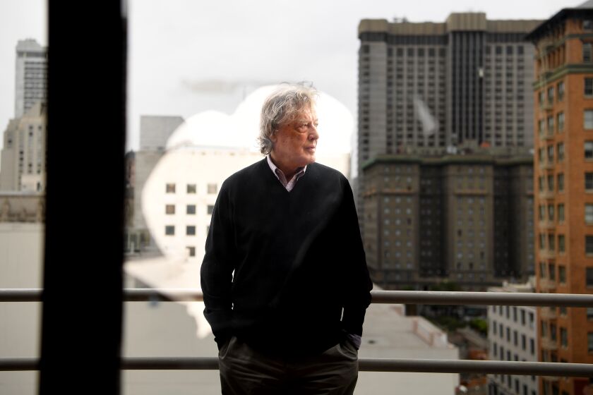 Tom Stoppard stands in front of a window looking out toward the San Francisco skyline.