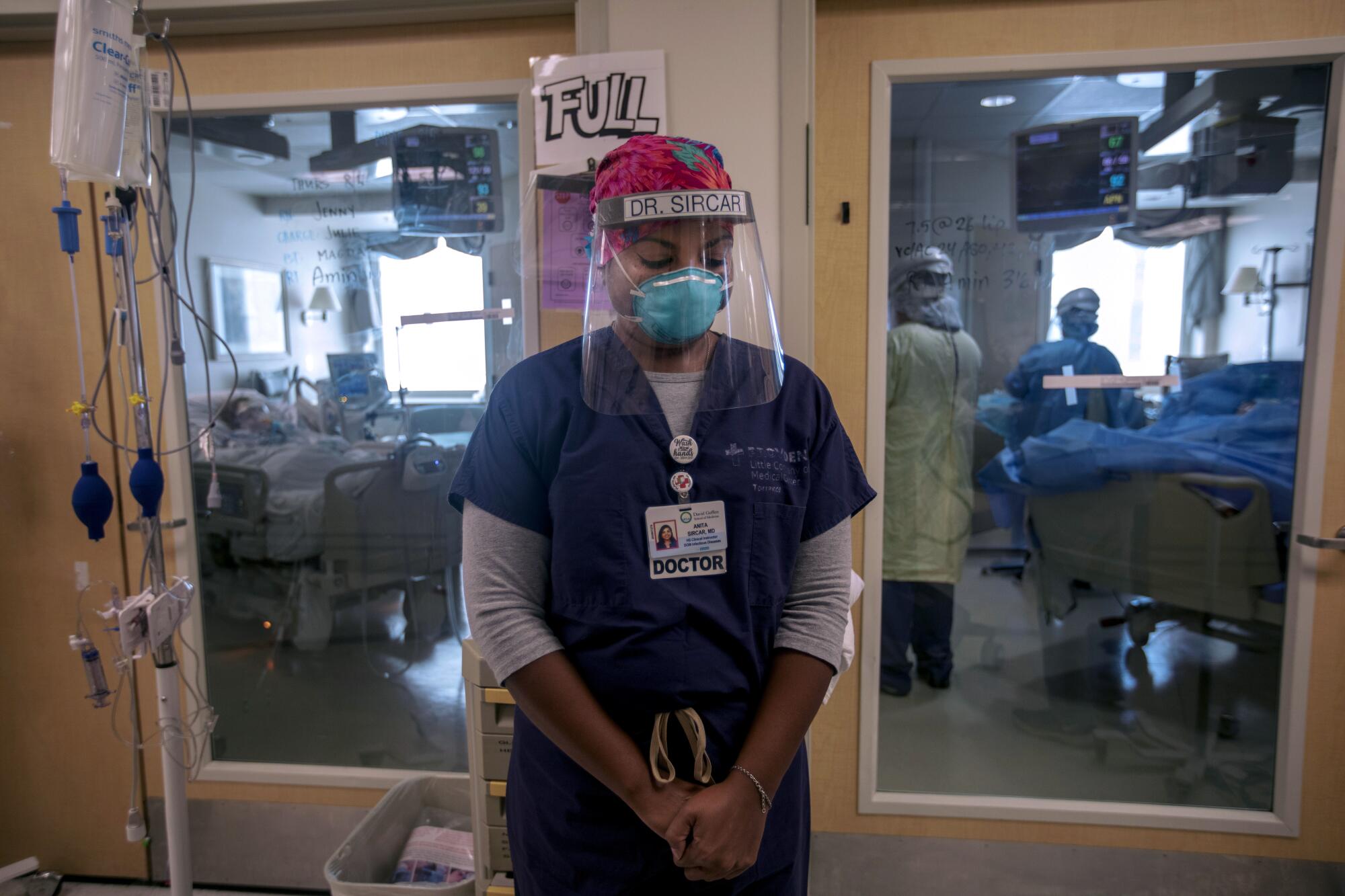 Dr. Anita Sircar bows her head outside a hospital room that has patients and hospital staff