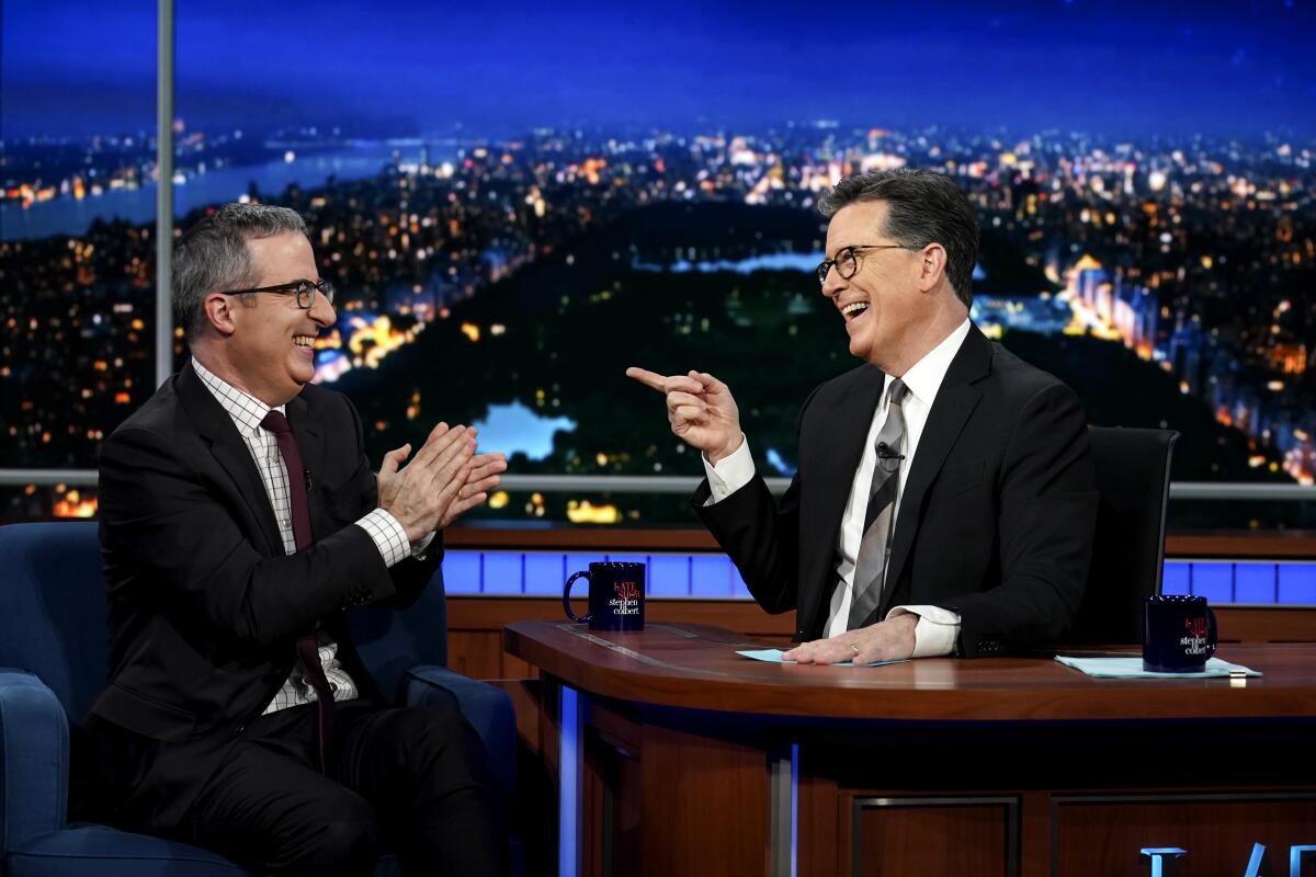 John Oliver sits in a chair and looks at Stephen Colbert, who is sitting behind a desk.