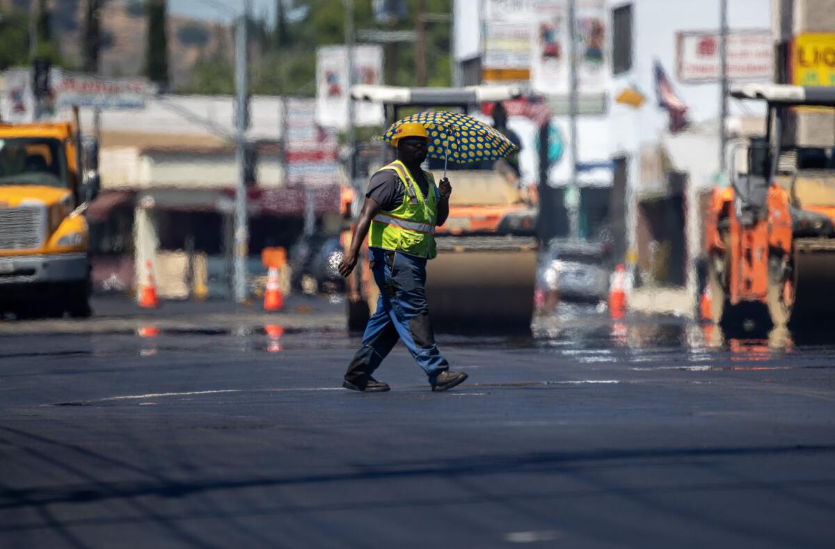 A worker carries an umbrella to ward off the sun as he walks over freshly paved asphalt. 