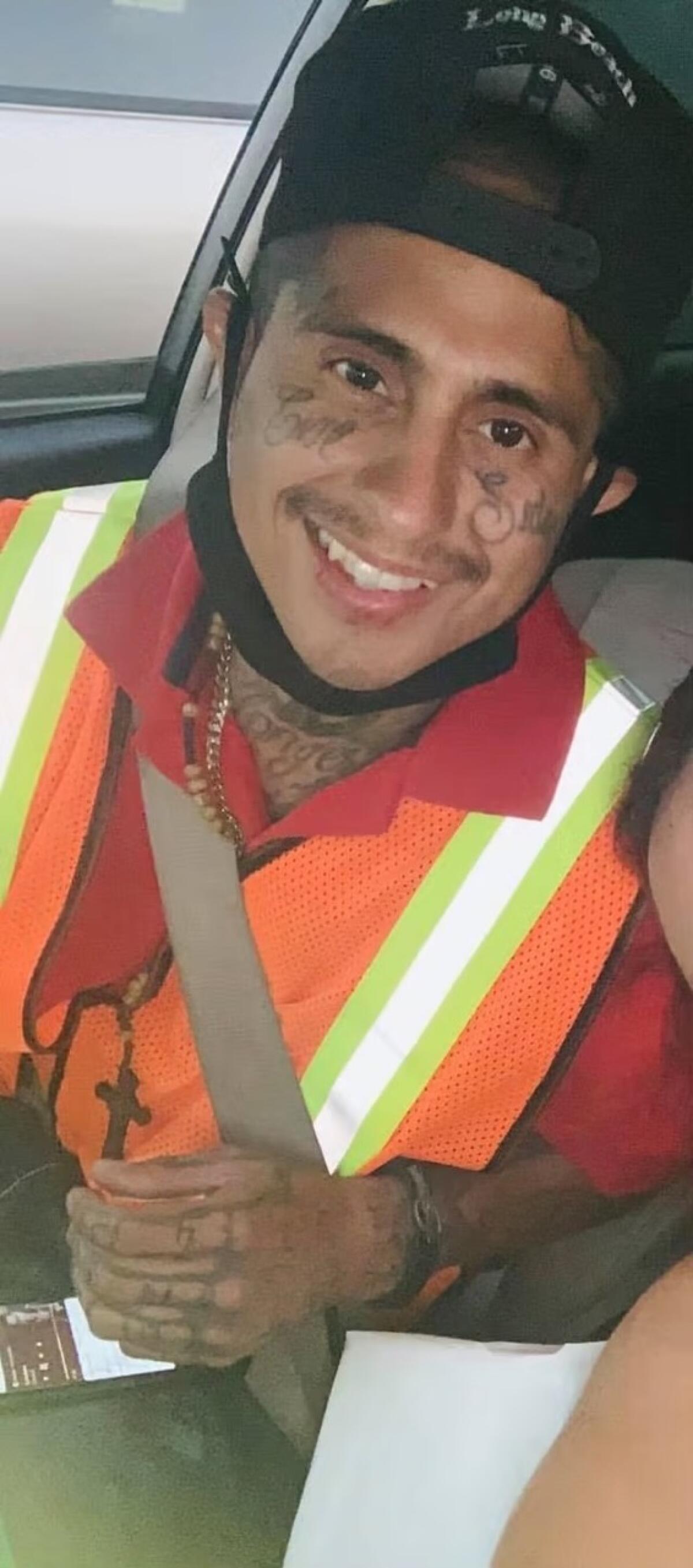 A man with tattoos wearing an orange high-visibility vest sits in a car