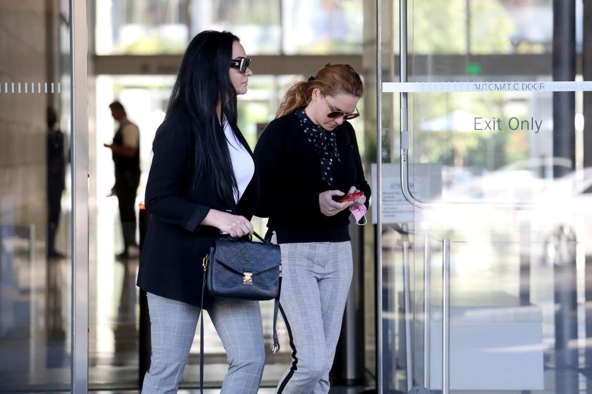 Two women, one looking down at her phone, walk near glass doors.