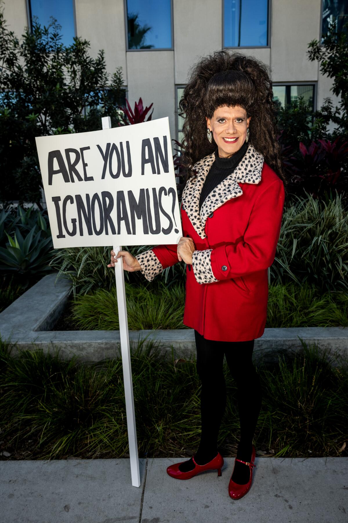 A man wearing a curly brunet wig, a red coat and red heels while smiling and holding a picket sign