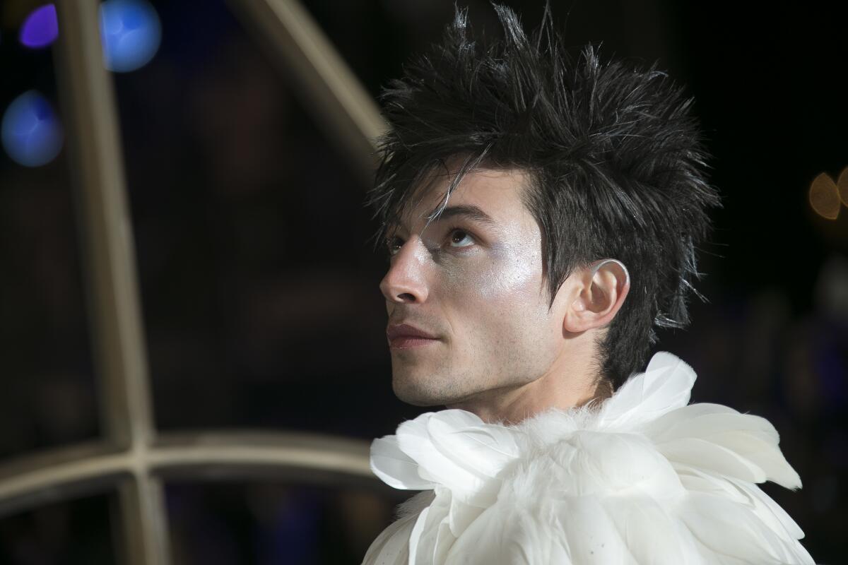 A person with black spiked hair wearing a feathery white collar looks up