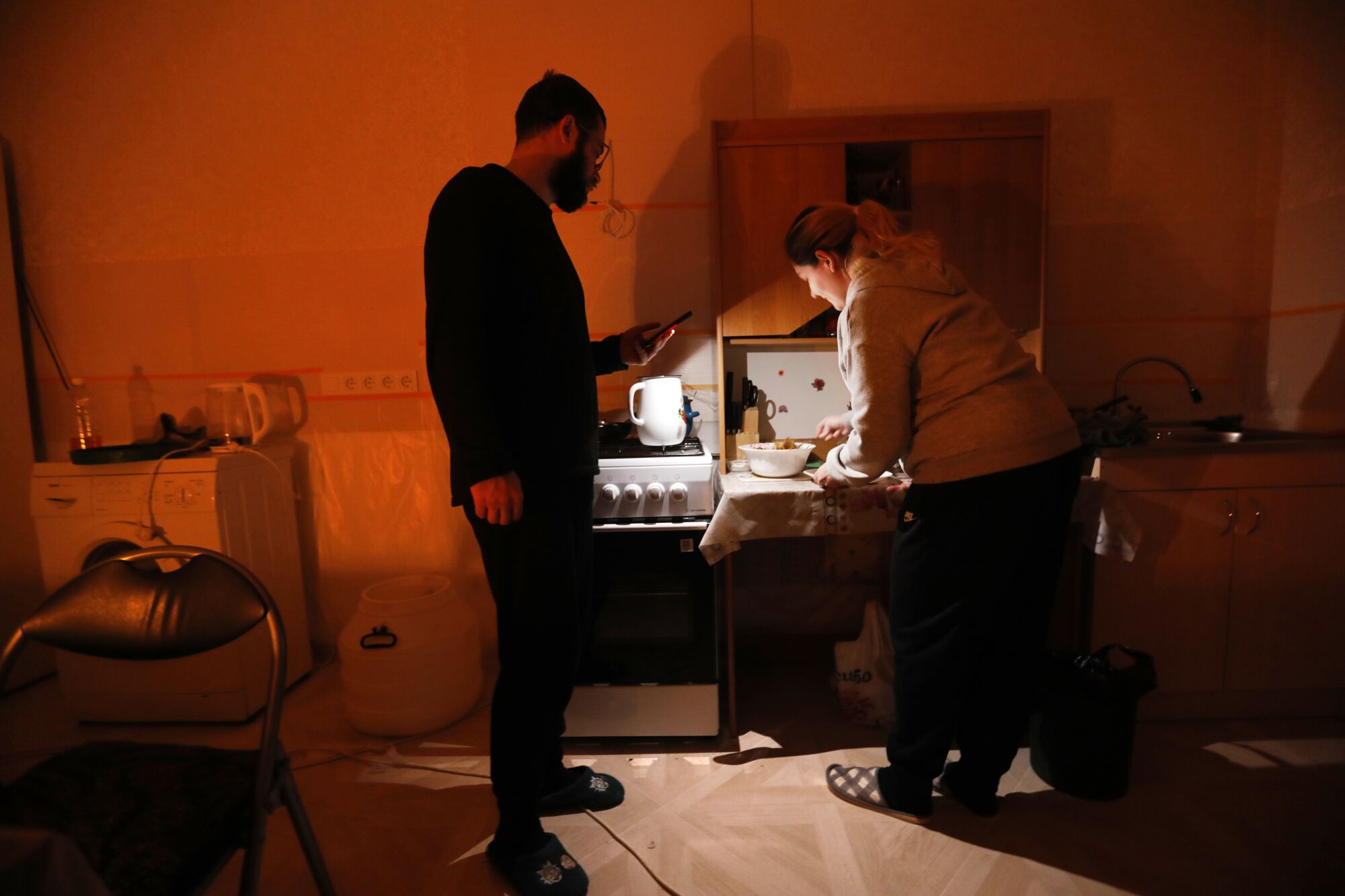 A man in dark clothing, left, uses his cellphone to provide light for a woman preparing food at a table