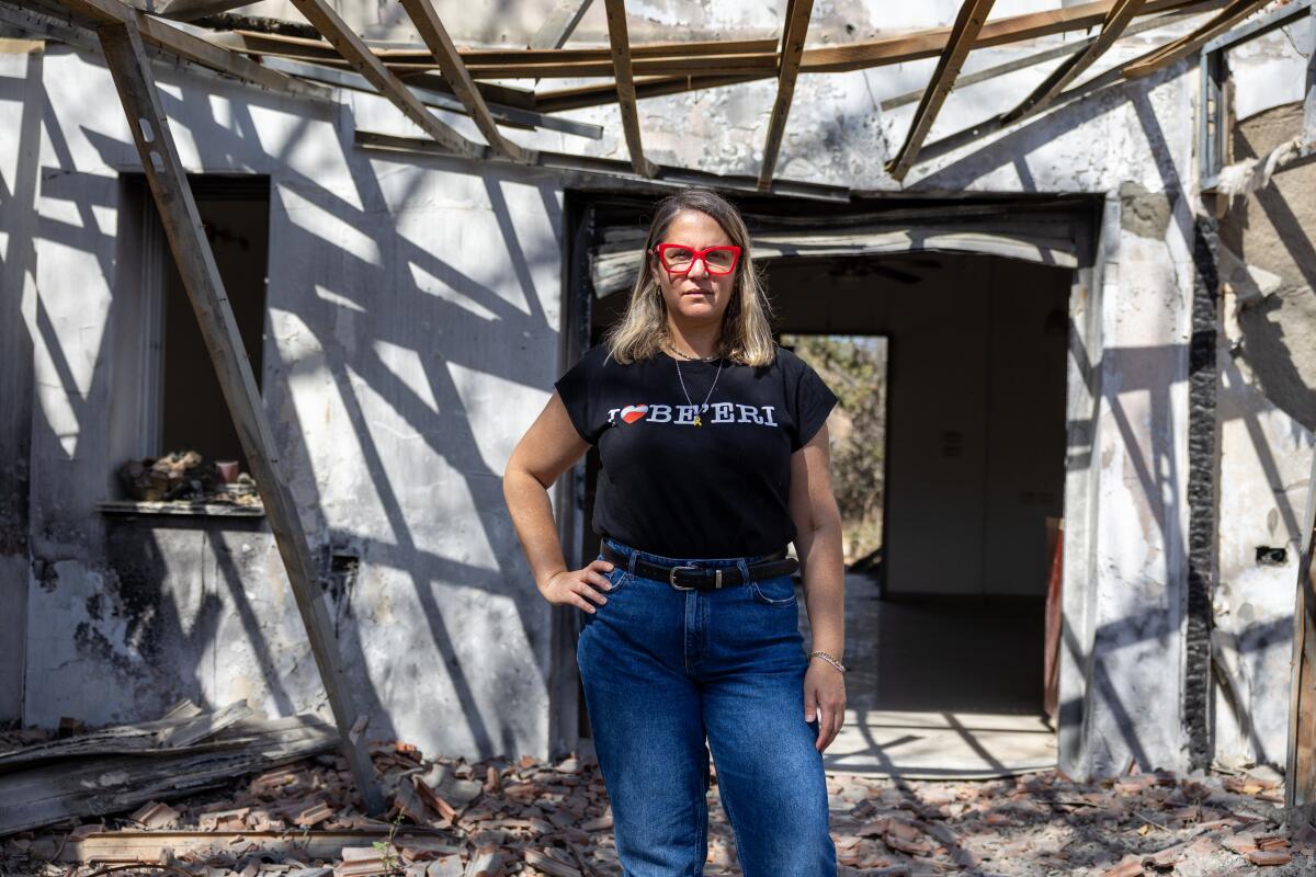 A woman with blond hair, in red glasses, black T-shirt and jeans, standing outside a white building with bare wood overhead