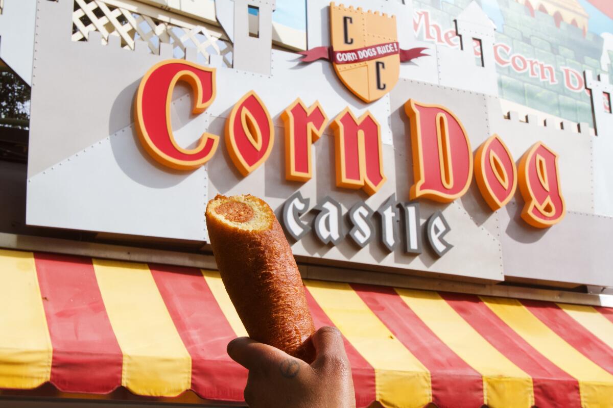 A hand holds up a corn dog in front of Corn Dog Castle's red-and-yellow signage.