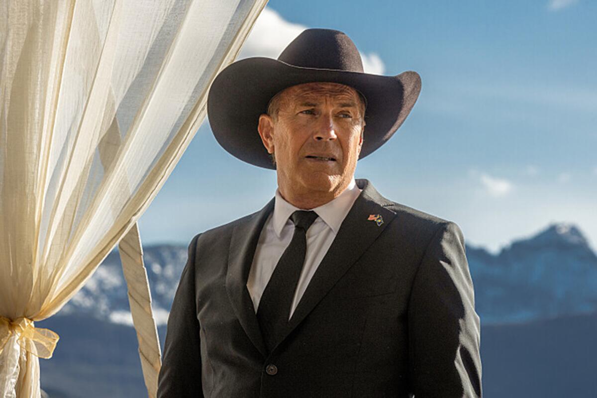 A man wears a cowboy hat and a suit and tie in front of a mountainous landscape