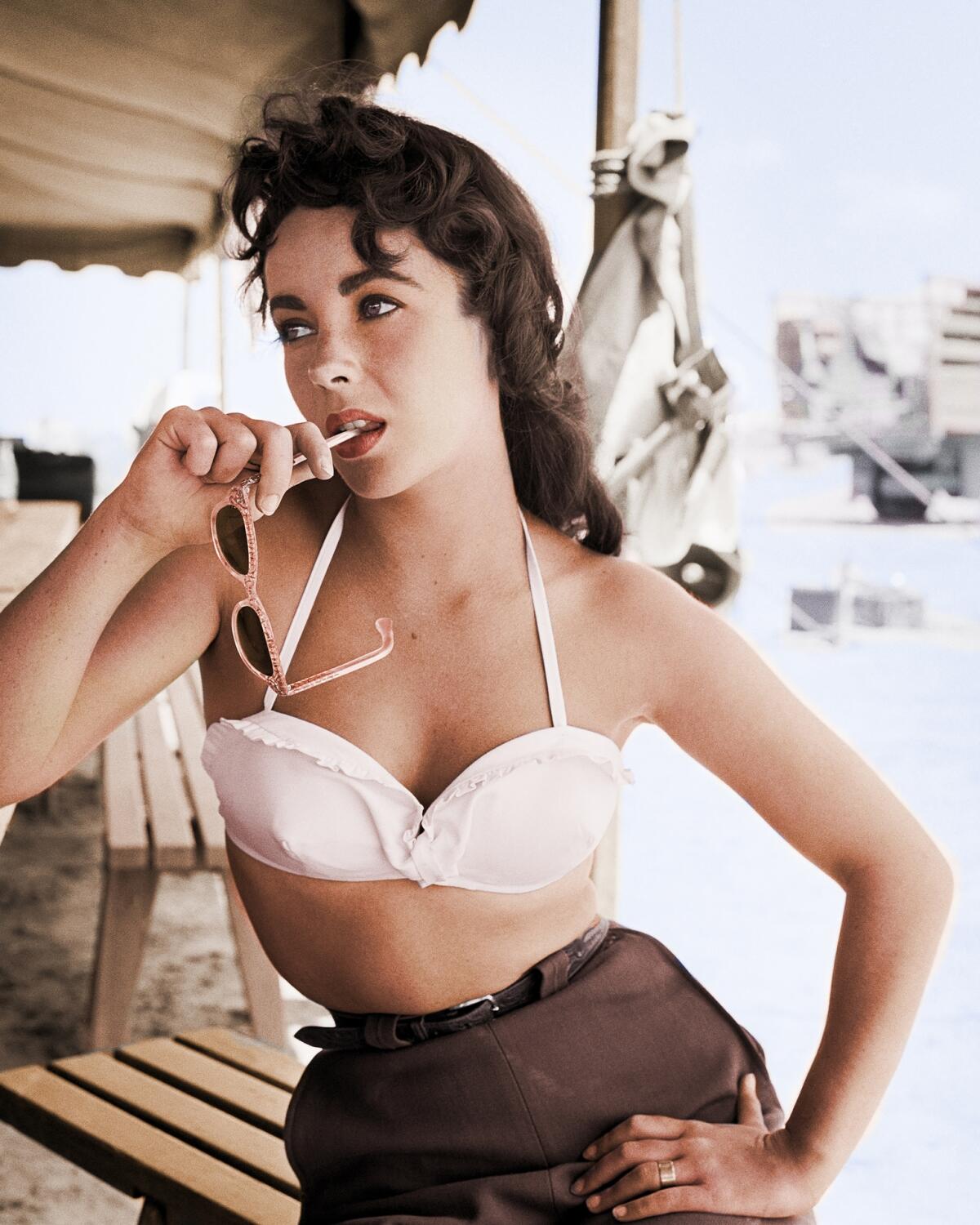 Elizabeth Taylor in a white bikini top and brown pants with a pair of sunglasses in her mouth.