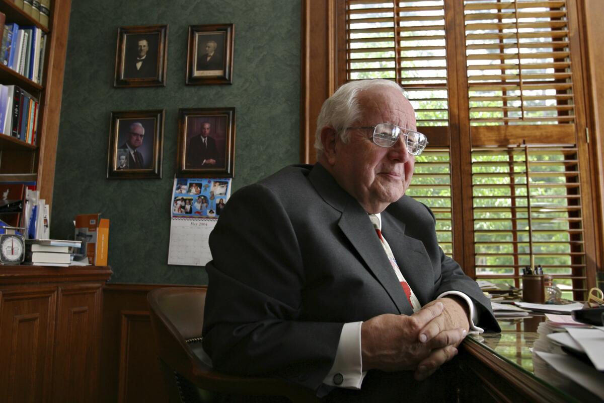 A man in a suit poses for a photo at a desk in his home.