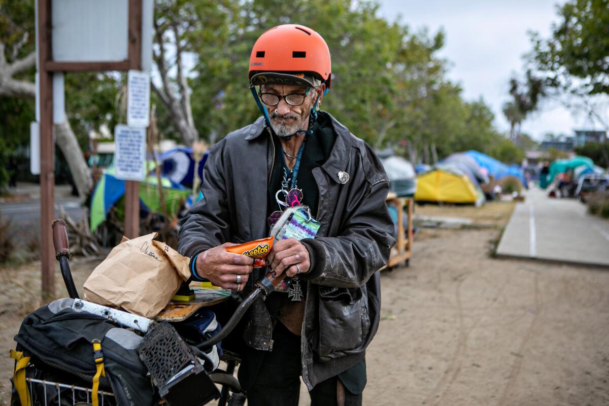 A man wearing a helmet eats Cheetos near a homeless encampment.
