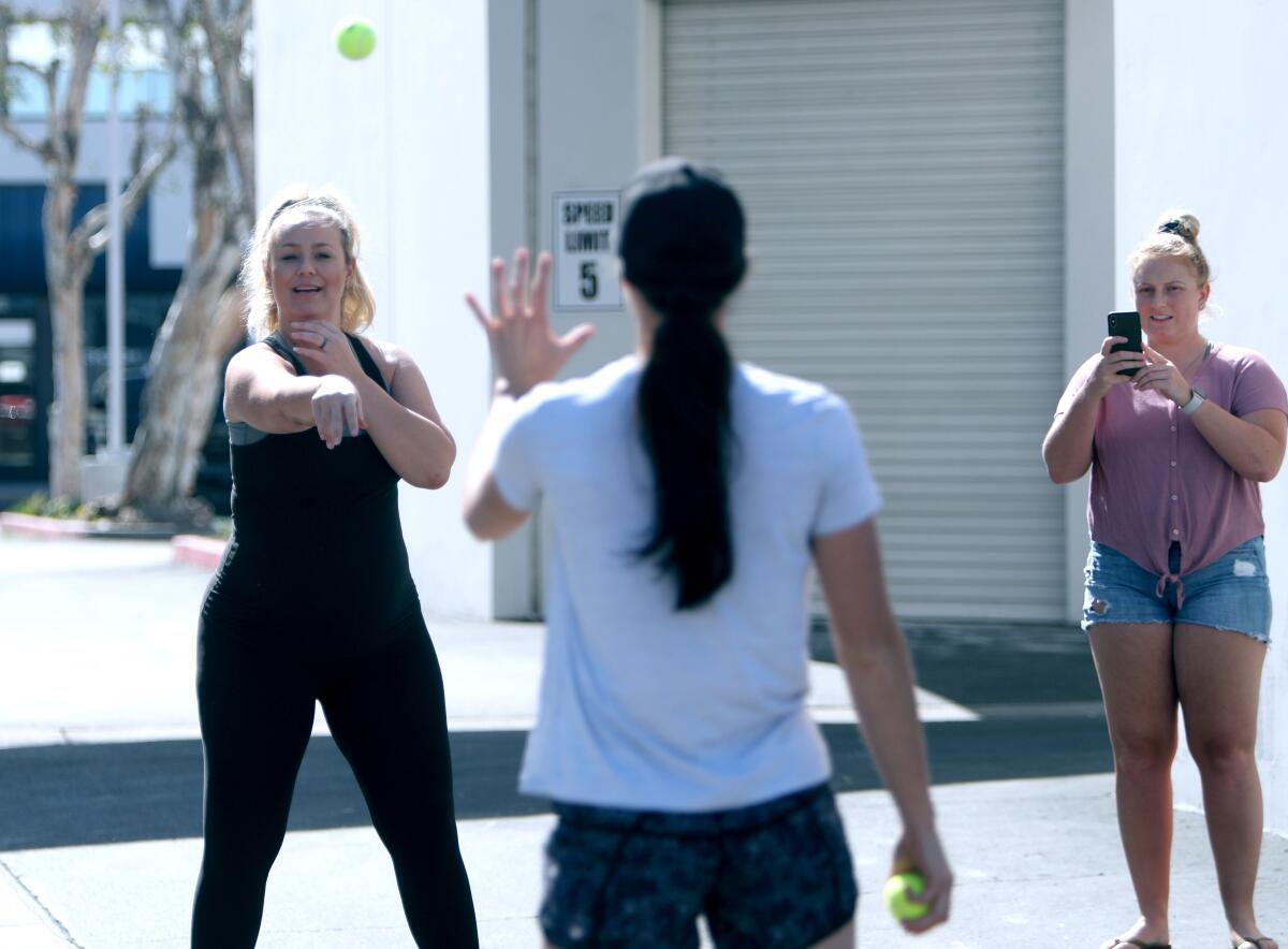 The Packaged Deal softball school co-founder Jen Schroeder, left, plays catch with co-founder Morgan Stuart, center, as Kayleen Shafer captures the action for hundreds of Facebook Live viewers outside her business in Anaheim on Tuesday. The class was for drills and skills softball students can do alone at home.